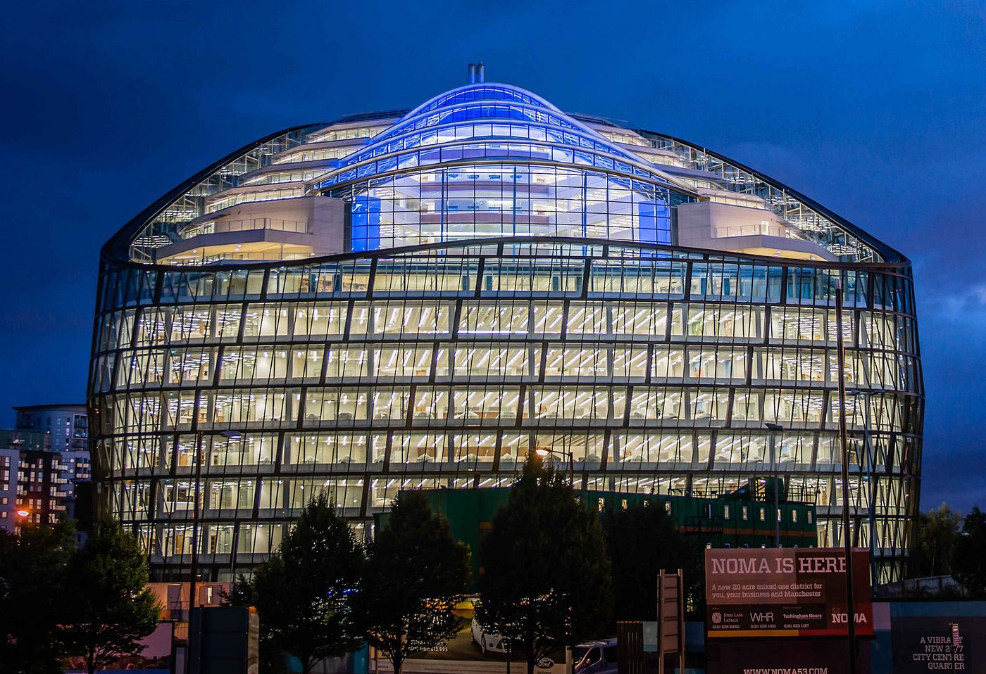 Foto: CO-OP Manchester: rundes, mehrgeschossiges Gebäude mit Glasfassade, vollständig von innen beleuchtet vor dunklem Nachthimmel