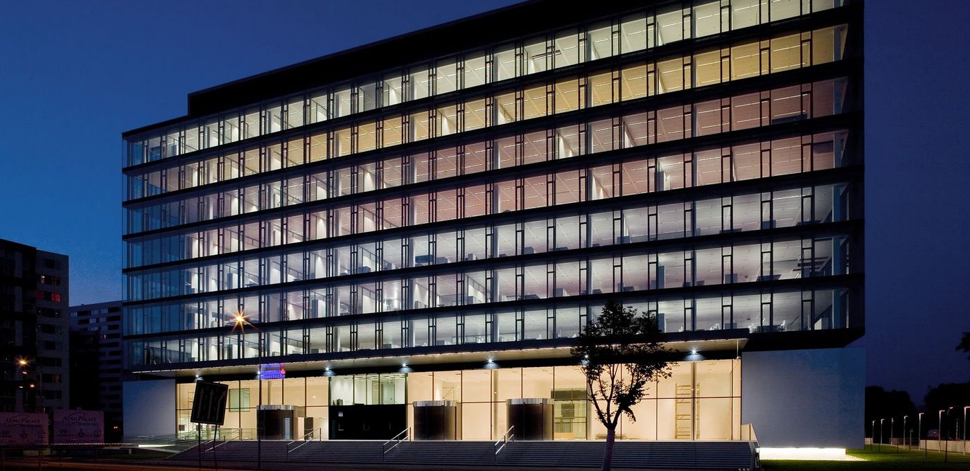 Foto: Bankgebäude SLSP: Nachtaufnahme eines mehrgeschossigen Gebäudes mit von innen in unterschiedlichen Farben beleuchteten Fenstern; eine Straße und Bäume im Vordergrund